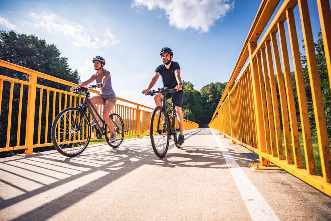 Zwei Fahrradfahrer auf einer Brücke mit gelben Geländer. Im Hintergrund ist der Himmel und der Wald zu sehen.